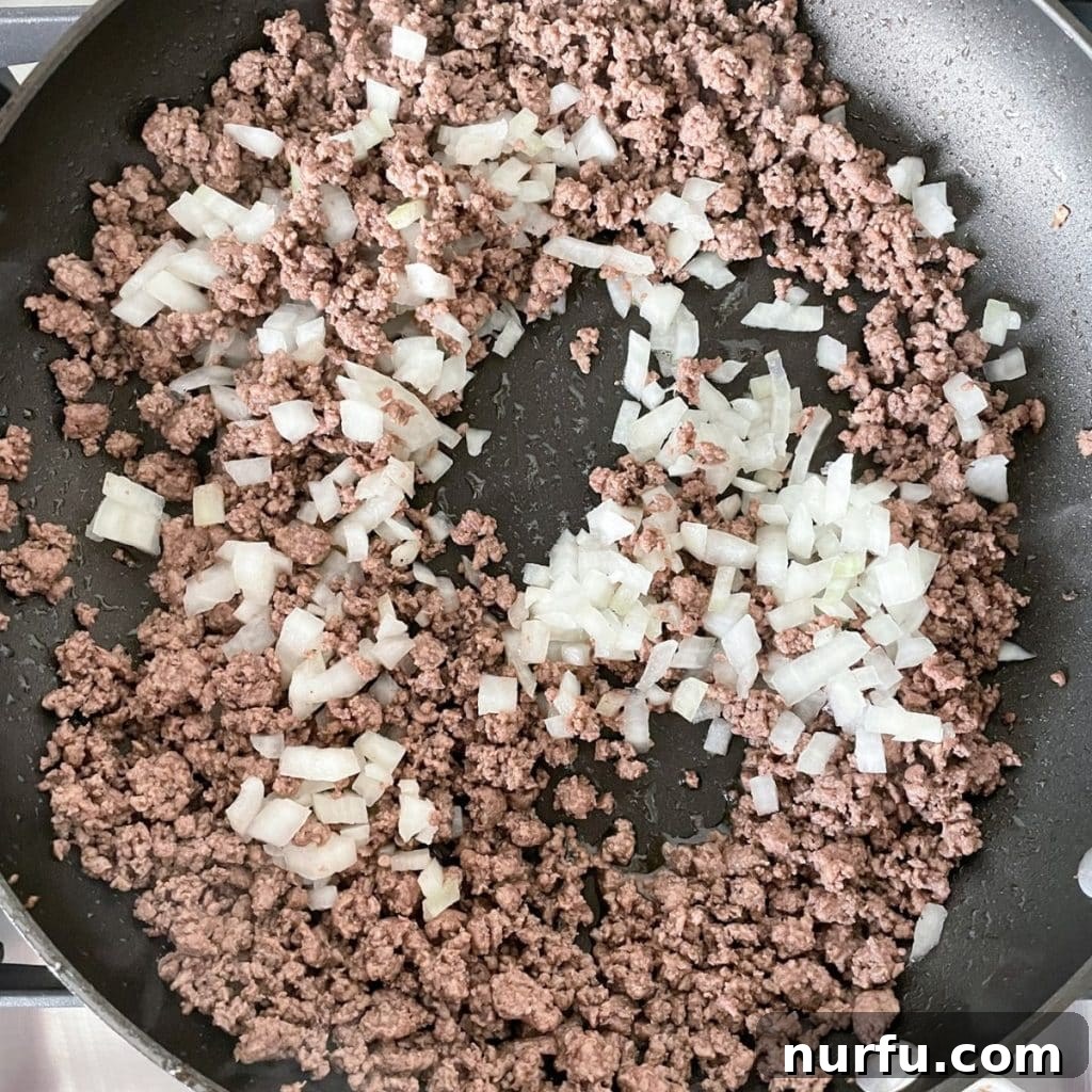 A large skillet showing browned ground beef mixed with softened diced onion, ready to be transferred for the next step of the pie preparation.