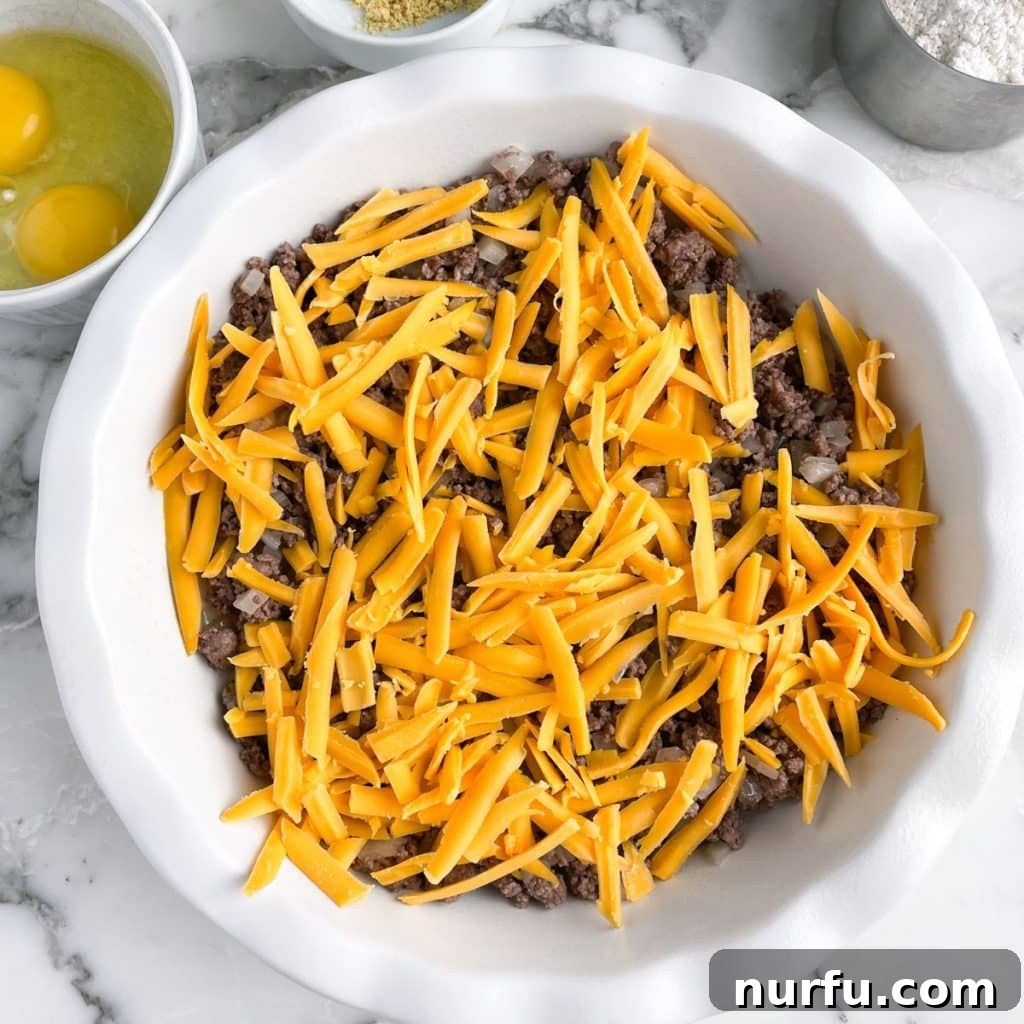 A pie plate filled with the savory ground beef and onion mixture, generously topped with shredded cheddar cheese, awaiting the Bisquick batter.