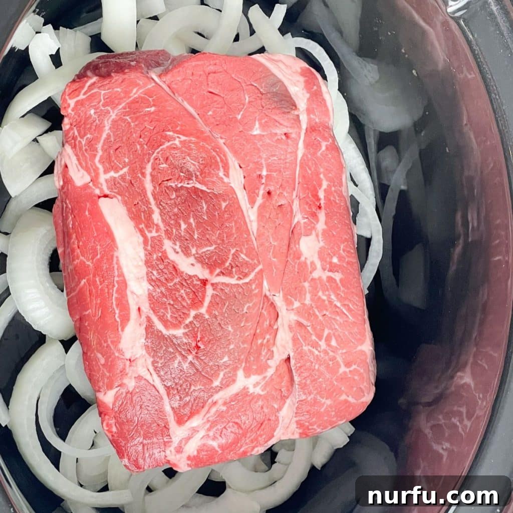 Melt-in-Your-Mouth Slow Cooker Beef 4 A large chuck roast sits atop a bed of sliced yellow onions inside a slow cooker pot, ready for the next steps of the recipe.