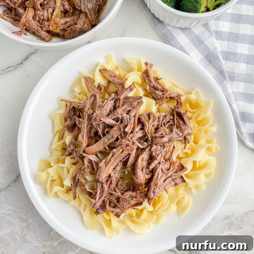 Melt-in-Your-Mouth Slow Cooker Beef 7 A generous serving of tender, flavorful shredded beef draped over a bed of golden egg noodles in a white bowl, garnished lightly with fresh herbs.