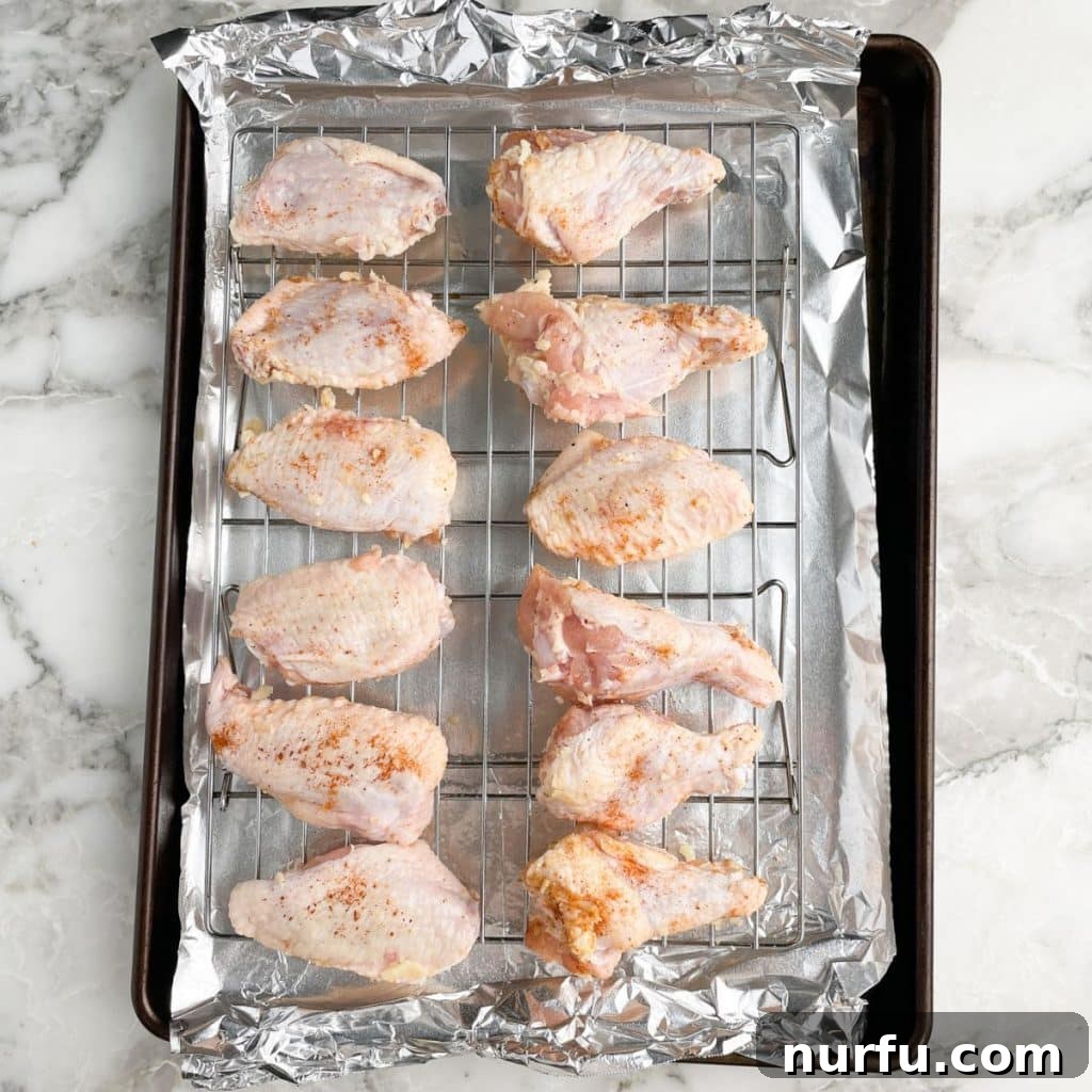 Uncooked, seasoned chicken wings neatly arranged in a single layer on a wire rack set over a baking sheet, precisely positioned for optimal oven baking.