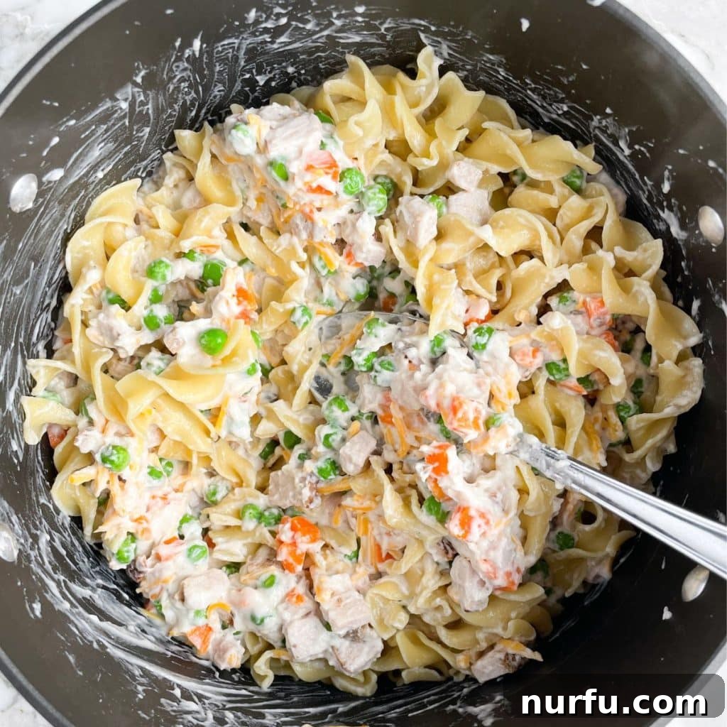 Hearty Pork Casserole 5 A close-up view of a pot filled with cooked egg noodles, vibrant peas and carrots, and a rich, creamy sauce, all mixed together.