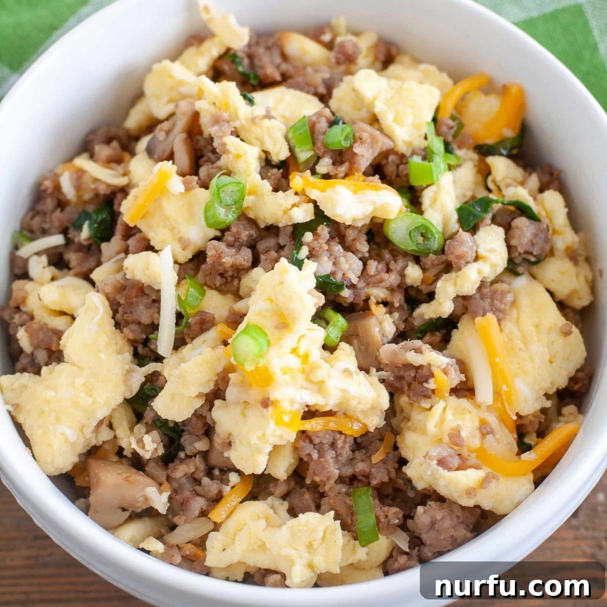Brinner Bonanza: 31 Recipes for Anytime 7 A white bowl brimming with a keto-friendly Egg and Sausage Bowl, featuring scrambled eggs, savory sausage, sautéed mushrooms, and vibrant spinach, topped with cheese.