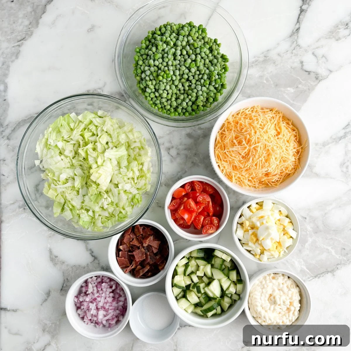 Layered Salad Perfection 3 An overhead view of various Seven Layer Salad ingredients laid out, including crisp lettuce, bright green peas, shredded cheese, diced cucumbers, creamy mayonnaise, red onion, and crumbled bacon, ready for assembly.