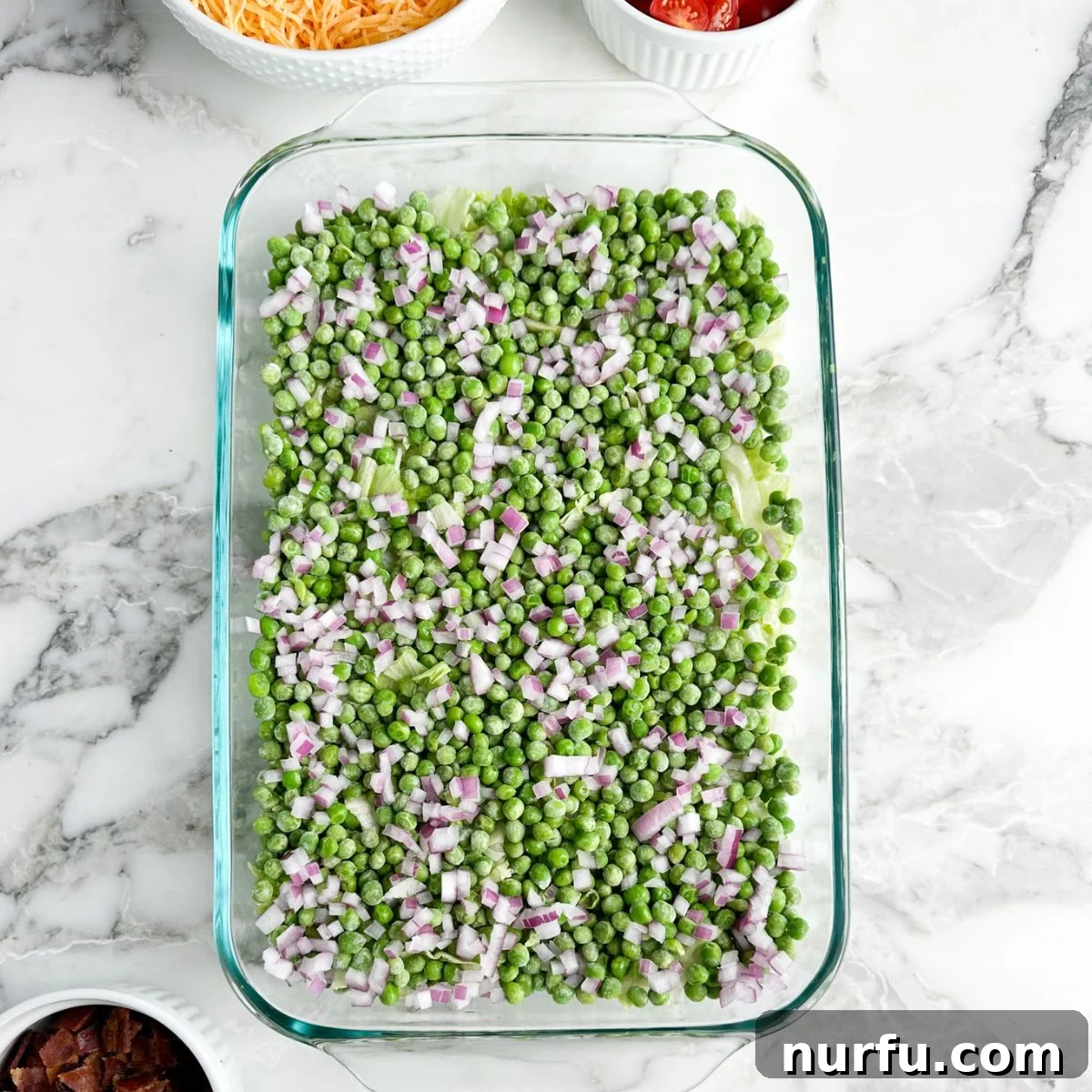 Layered Salad Perfection 4 A casserole dish with the first layers of Seven Layer Salad: a base of shredded lettuce topped with bright green peas and finely diced red onions, ready for the next ingredients.