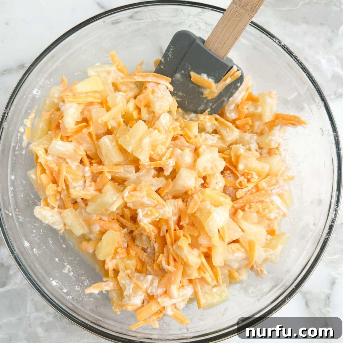 Tropical Pineapple Delight 4 A large mixing bowl containing a creamy mixture of pineapple tidbits and shredded cheddar cheese, with a spatula poised to continue mixing, illustrating the casserole filling preparation.