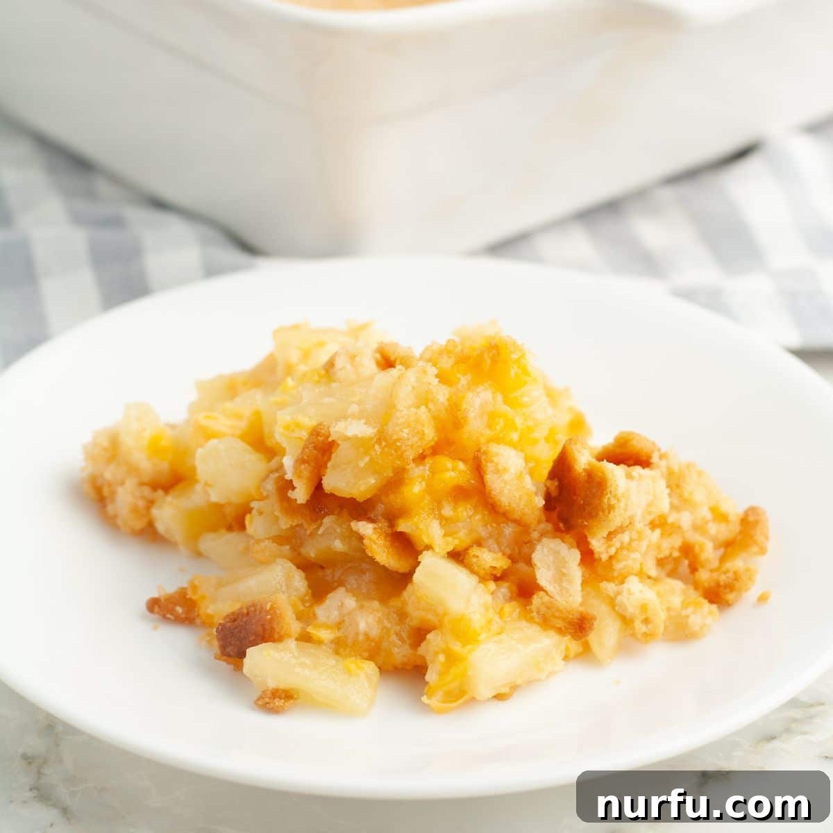Tropical Pineapple Delight 7 A perfectly portioned slice of warm pineapple casserole resting on a pristine white plate, showcasing its appealing golden, crisp topping and the rich, cheesy pineapple filling nestled underneath.