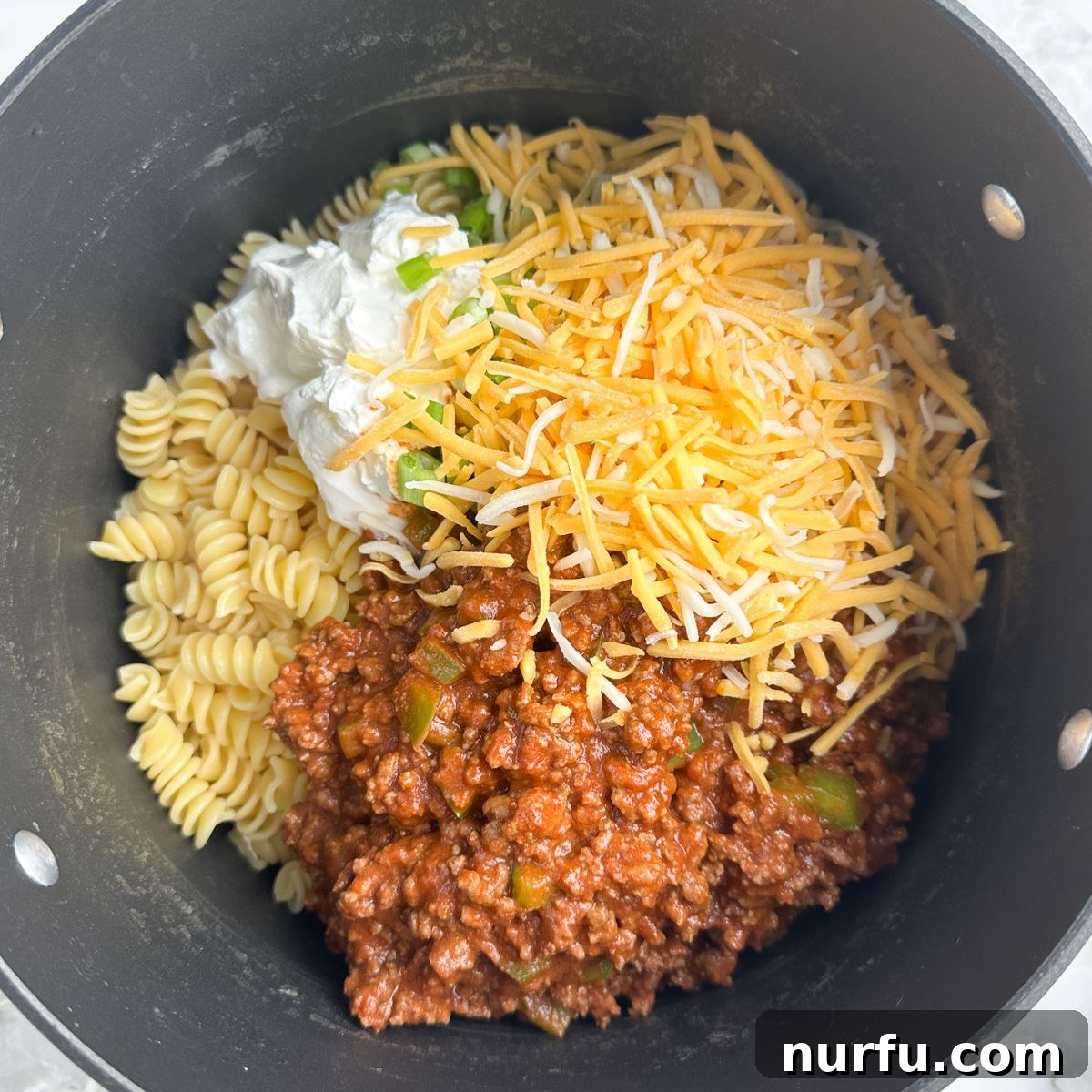 A large pot brimming with cooked pasta, seasoned ground beef, melted shredded cheese, and a dollop of sour cream, ready to be mixed into a creamy, savory Taco Twist Casserole.