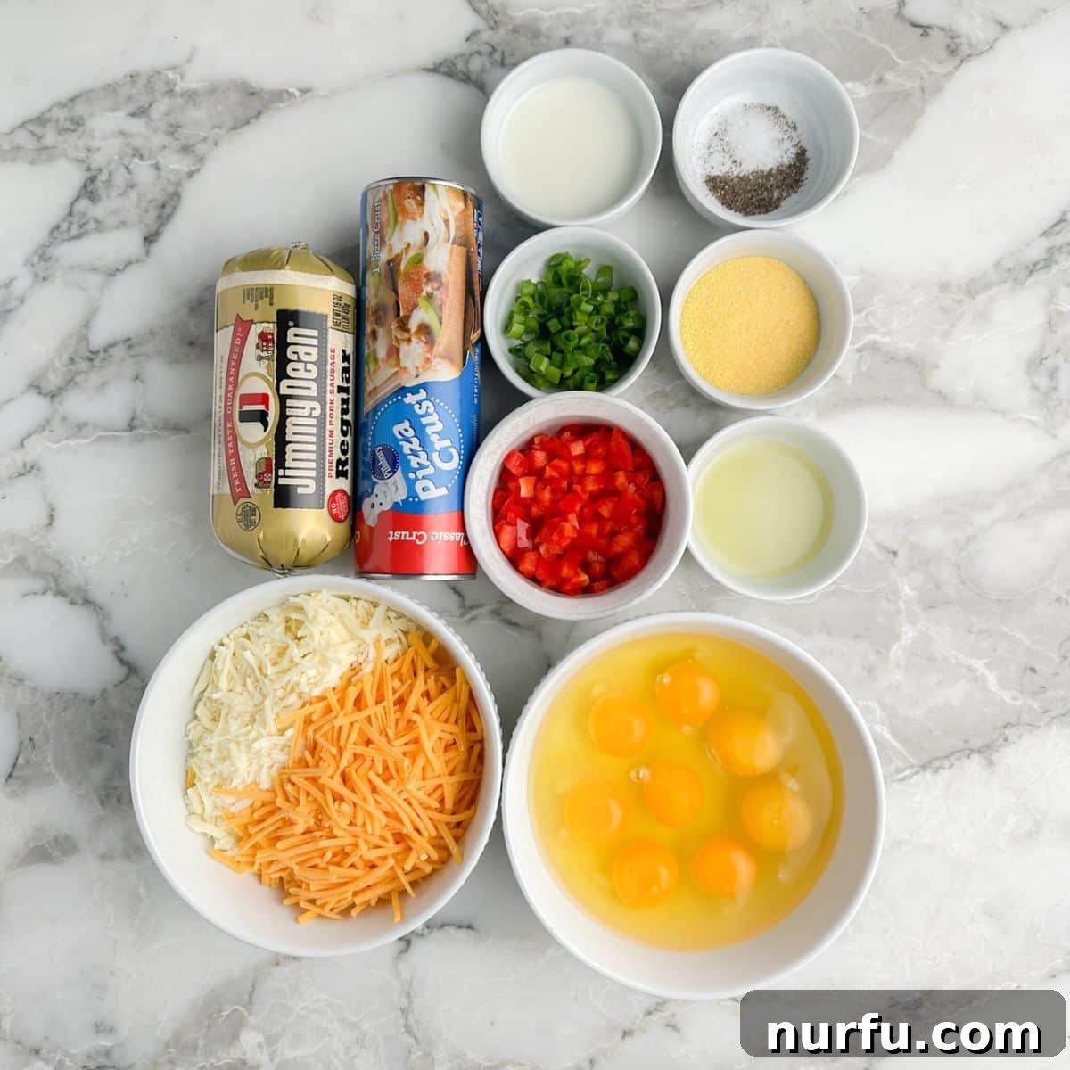 A collection of fresh breakfast pizza ingredients laid out on a table: a bowl of shredded cheese, a roll of pizza dough, cooked sausage, whisked eggs, diced bell pepper, and chopped green onion, ready for assembly.