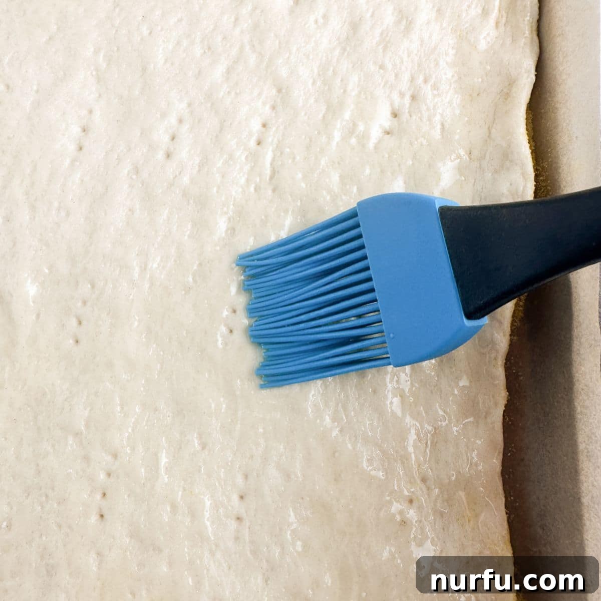 A close-up of pizza dough on a baking sheet, with olive oil being brushed onto its surface before baking.