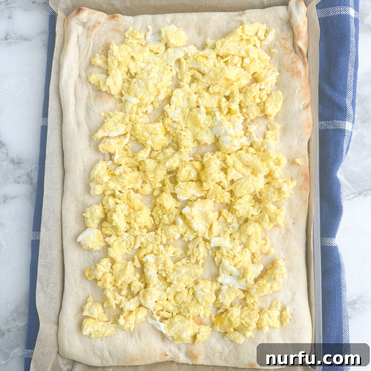 A partially baked pizza crust covered with a generous layer of fluffy scrambled eggs, ready for further toppings.