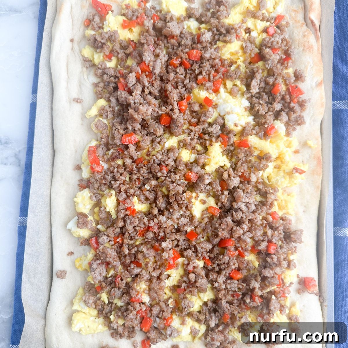 A close-up of a pizza crust topped with scrambled eggs and evenly distributed crumbled sausage, ready for cheese.