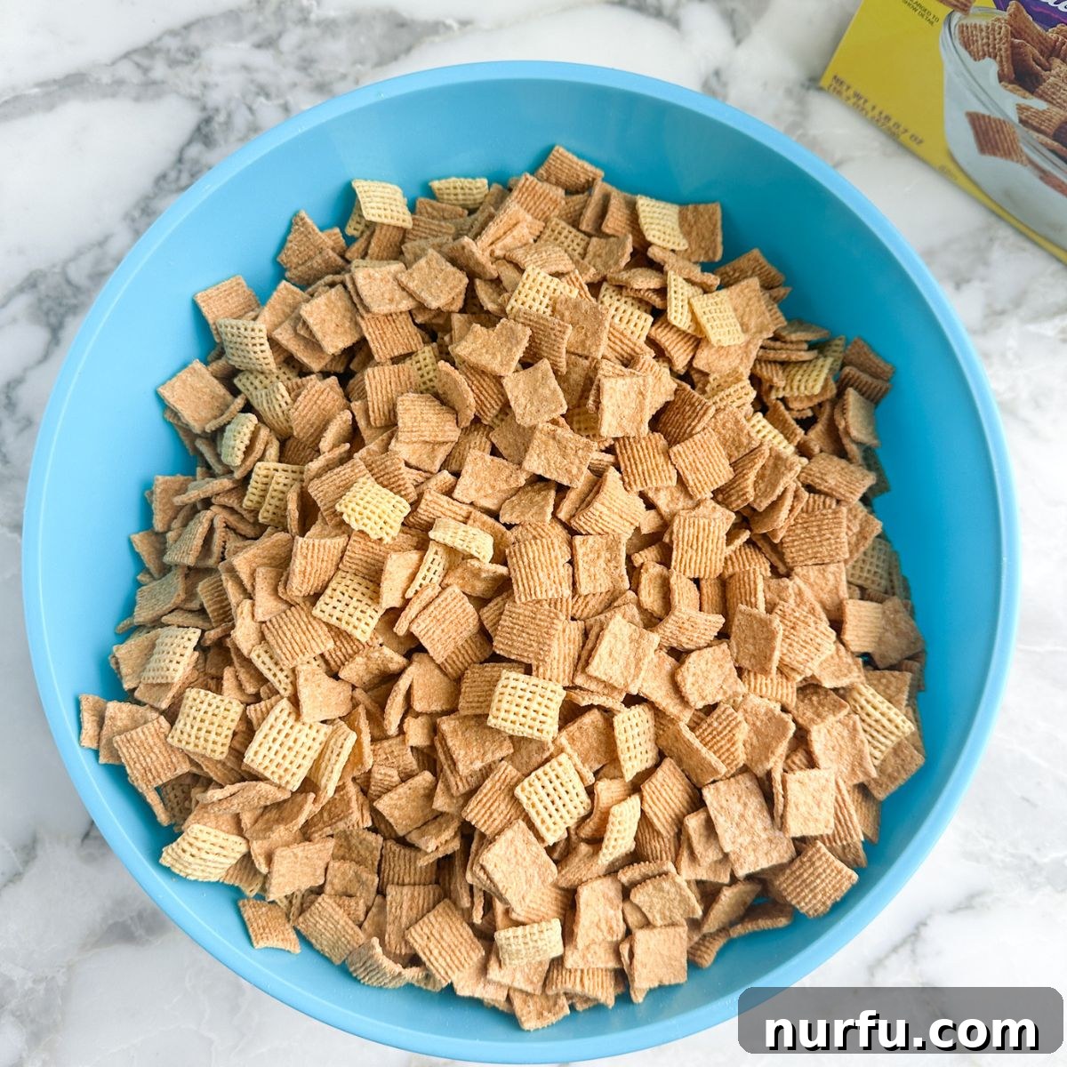 A vibrant blue mixing bowl filled with a blend of golden Golden Grahams cereal and square-shaped Corn Chex cereal, perfectly prepared for the next step in making Sweet Chex Mix.