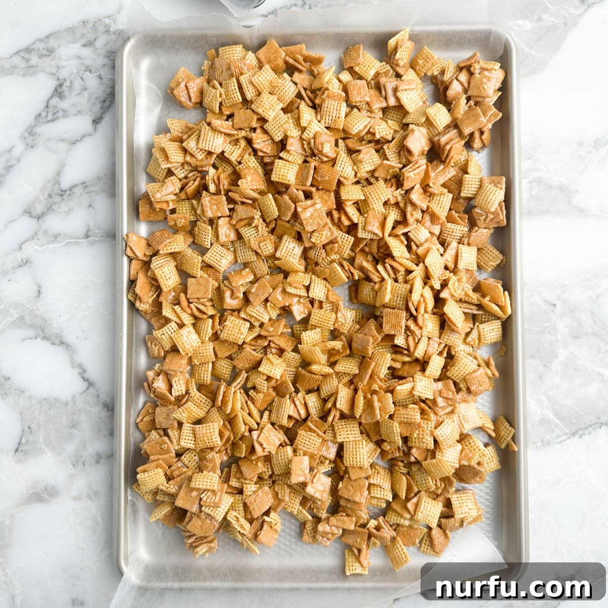 A baking sheet covered with parchment paper, laden with the fresh, still-warm Sweet Chex Mix, spread out to cool and harden.