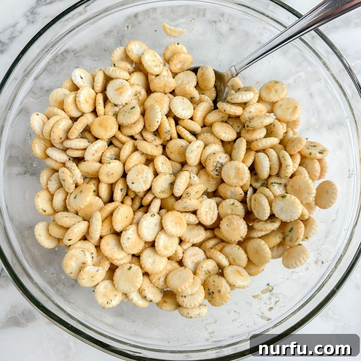 Homemade Savory Oyster Crackers in Minutes 5 Oyster crackers in a glass bowl with a spoon, being tossed with seasoning.