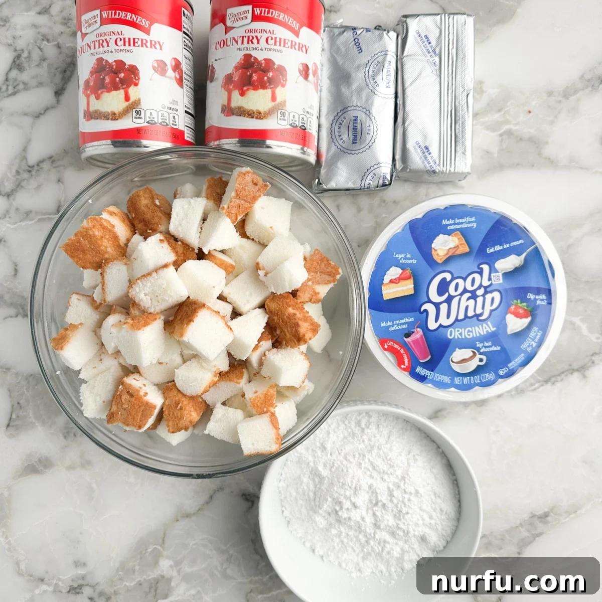 A collection of ingredients for Cherries in the Snow, including cans of cherry pie filling, cubed angel food cake, softened cream cheese, powdered sugar, and thawed whipped topping.