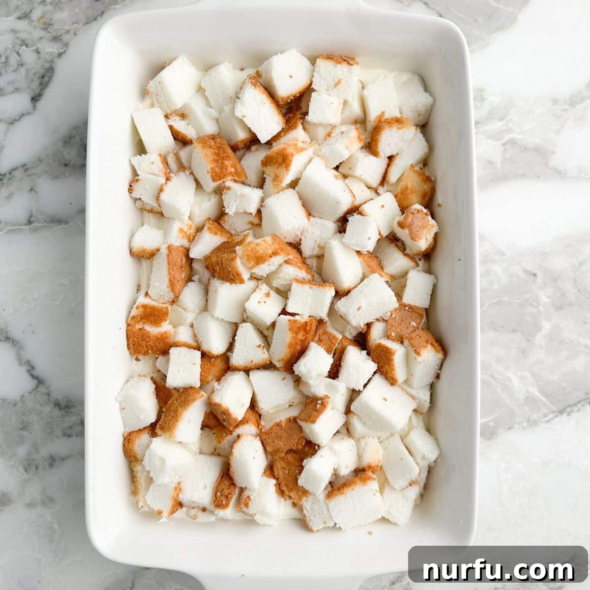 A 9x13 casserole dish with a layer of cubed angel food cake neatly arranged over the creamy base, ready for the cherry topping.