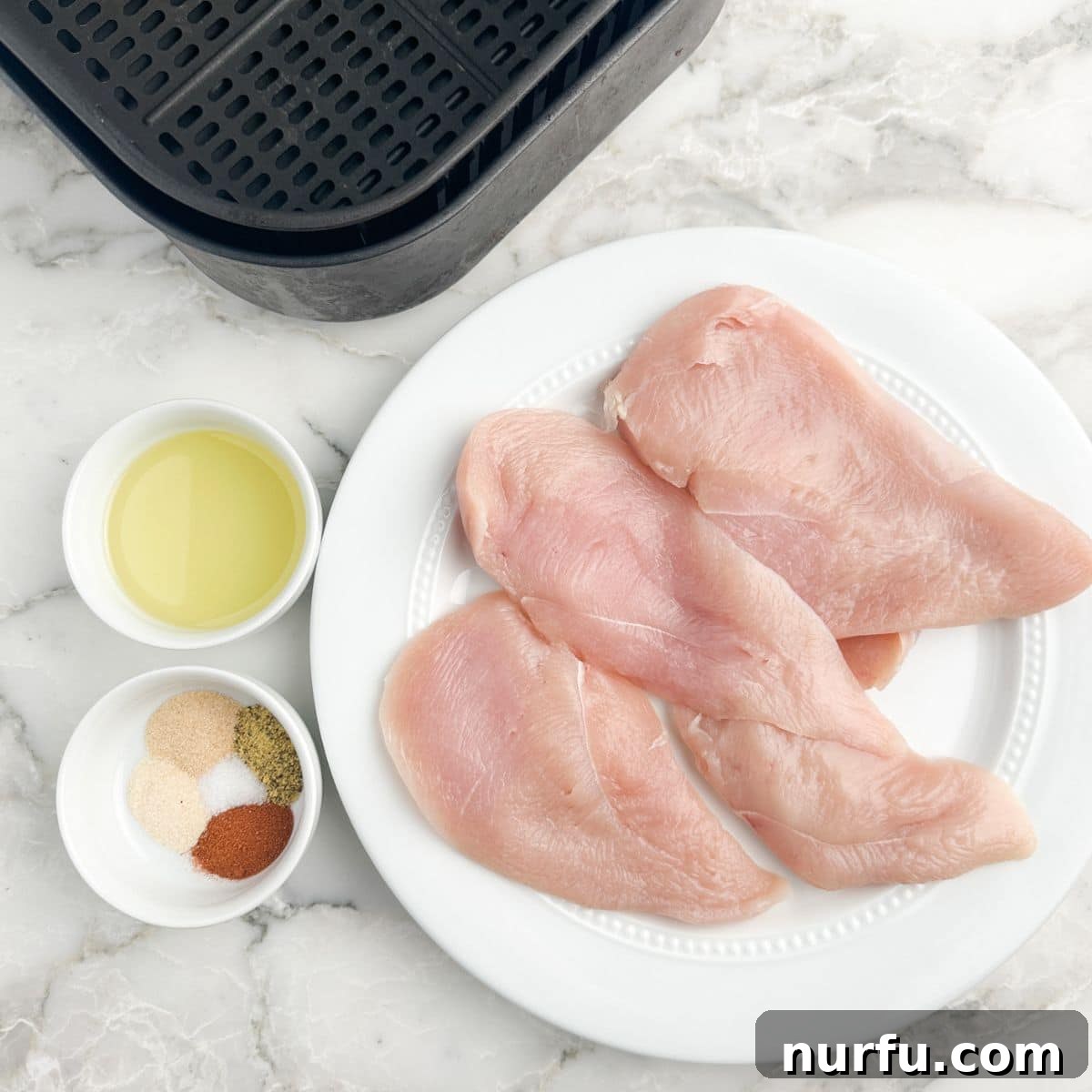 Quick Air Fryer Thin Chicken Breasts 3 Thin chicken breasts on a white plate, alongside a small bowl of mixed seasonings and a bottle of olive oil, ready for preparation.
