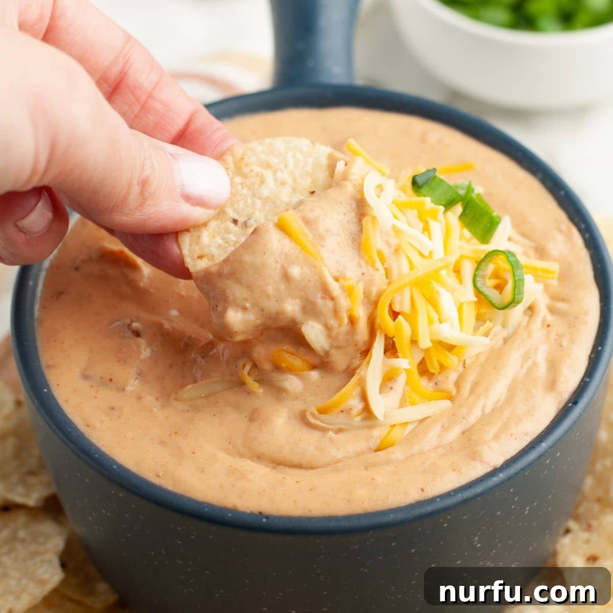 Creamy Fiesta Bean Dip 2 A vibrant bowl of refried bean dip, garnished with fresh cilantro, with a tortilla chip poised for dipping. The dip looks creamy and appealing, set against a warm, inviting backdrop.