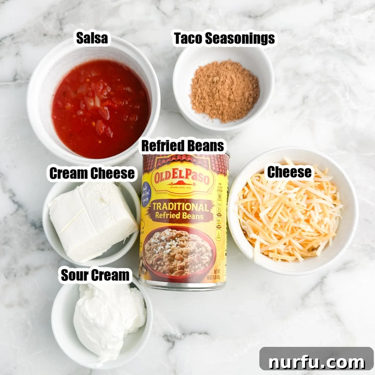 Creamy Fiesta Bean Dip 3 A flat lay photograph showcasing all the ingredients required for the refried bean dip: a can of refried beans, a block of cream cheese, a bowl of sour cream, taco seasoning, salsa, and a mound of shredded cheese, neatly arranged on a rustic surface.