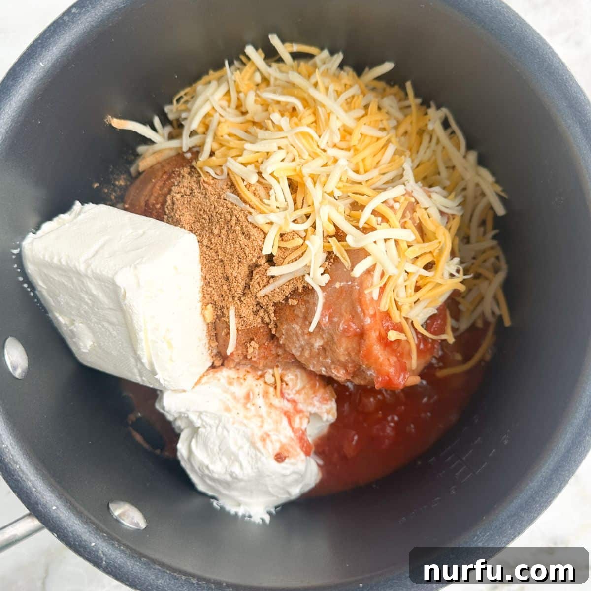 Creamy Fiesta Bean Dip 4 A medium saucepan on a stove top, filled with a mixture of refried beans, cream cheese, sour cream, and shredded cheese before being stirred. The ingredients are visibly distinct.