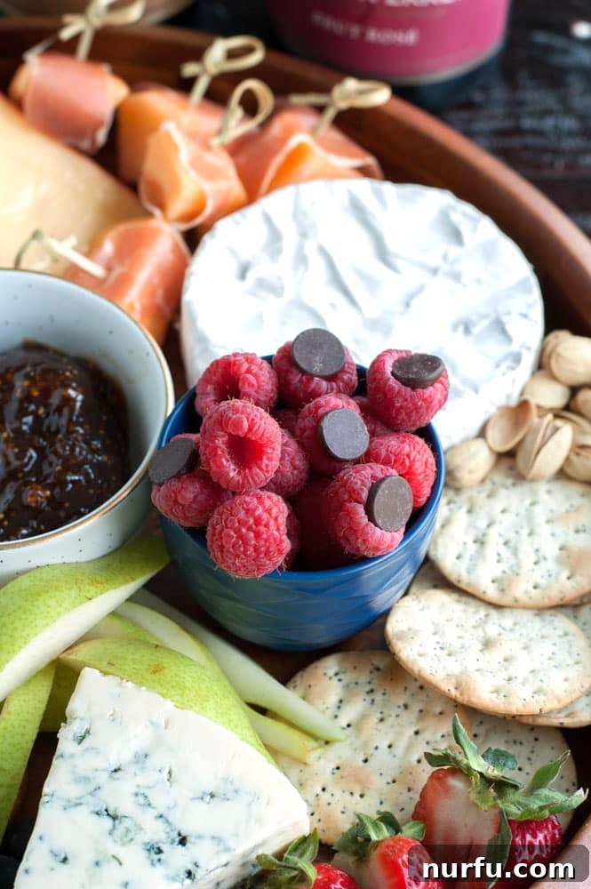 A dessert-focused section of a cheese board, featuring a bowl of raspberries with dark chocolate chips, crackers, and cheese.