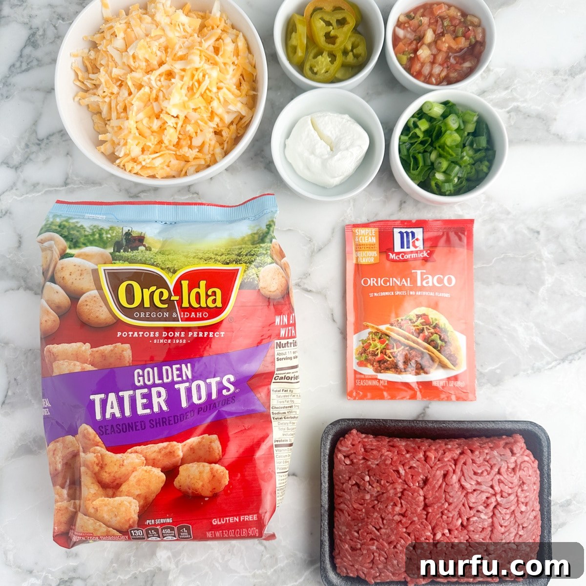 Ultimate Beef Tater Tot Nachos 3 A flat lay of ingredients for totchos including a bag of tater tots, a bowl of shredded cheese, raw ground beef, taco seasoning, sour cream, salsa, and diced green onions.