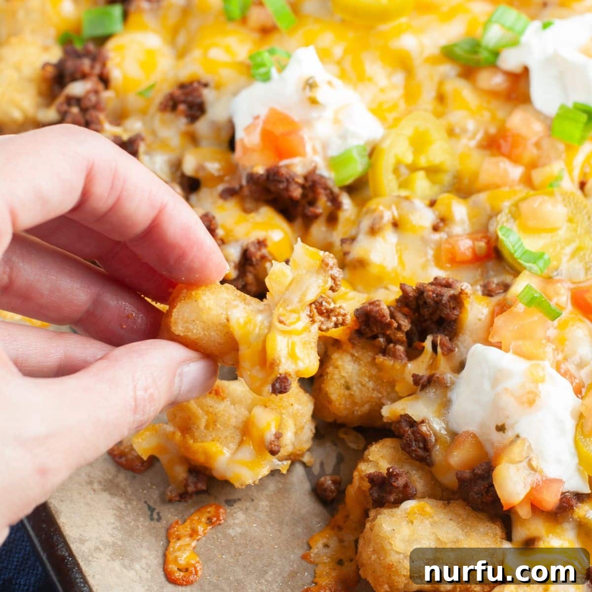 Ultimate Beef Tater Tot Nachos 5 A hand holding a serving of ground beef totchos, showing the crispy tots, seasoned beef, and melted cheese with fresh toppings.