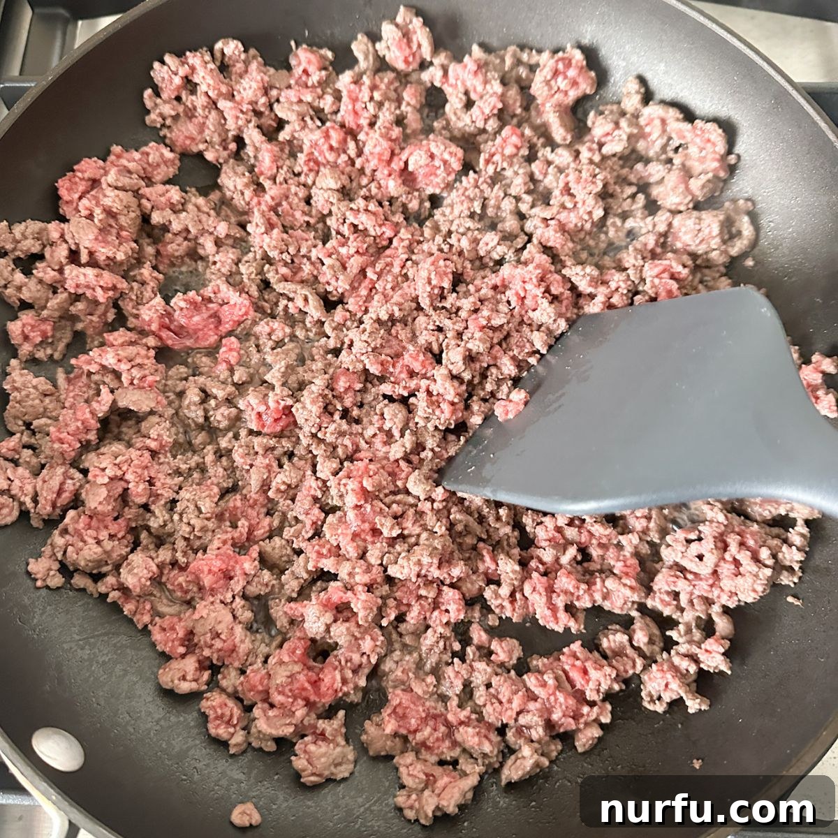 Seasoning Secrets for Ground Beef 5 Ground beef cooking in a large skillet over medium heat, being skillfully broken apart into smaller pieces with a wooden spoon, releasing steam as it browns.