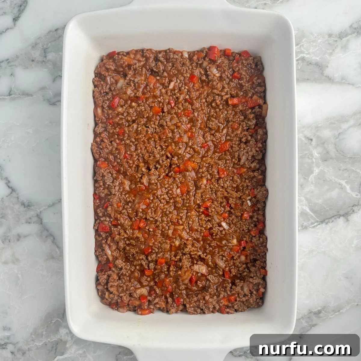 A 9x13 inch casserole dish filled with the evenly spread sloppy joe meat mixture, ready for the next layers.
