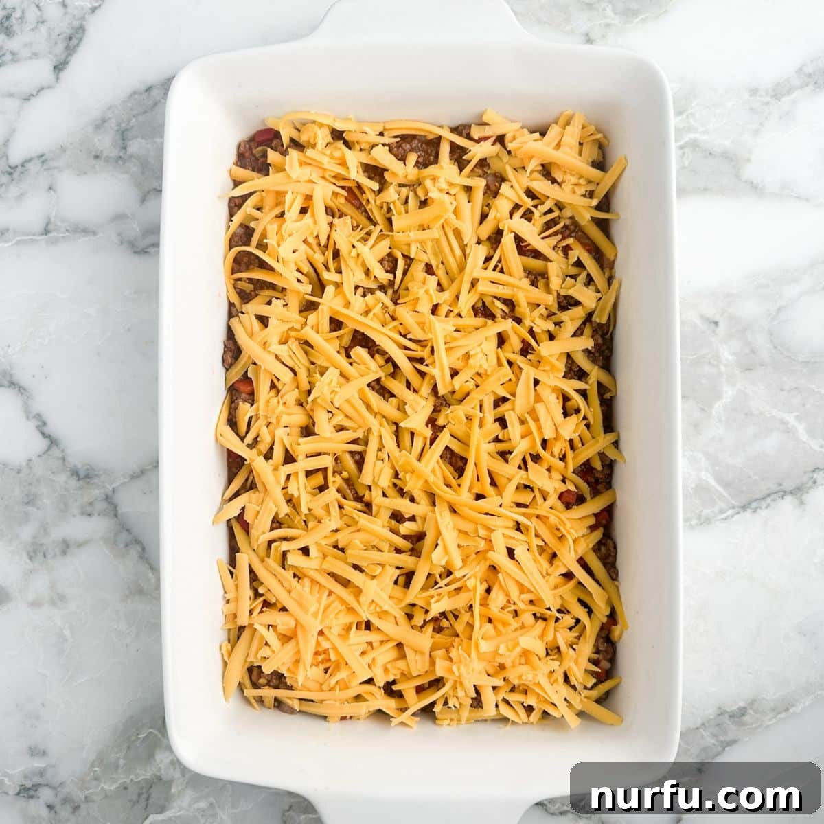 A casserole dish with the sloppy joe beef mixture generously topped with a layer of shredded cheddar cheese.