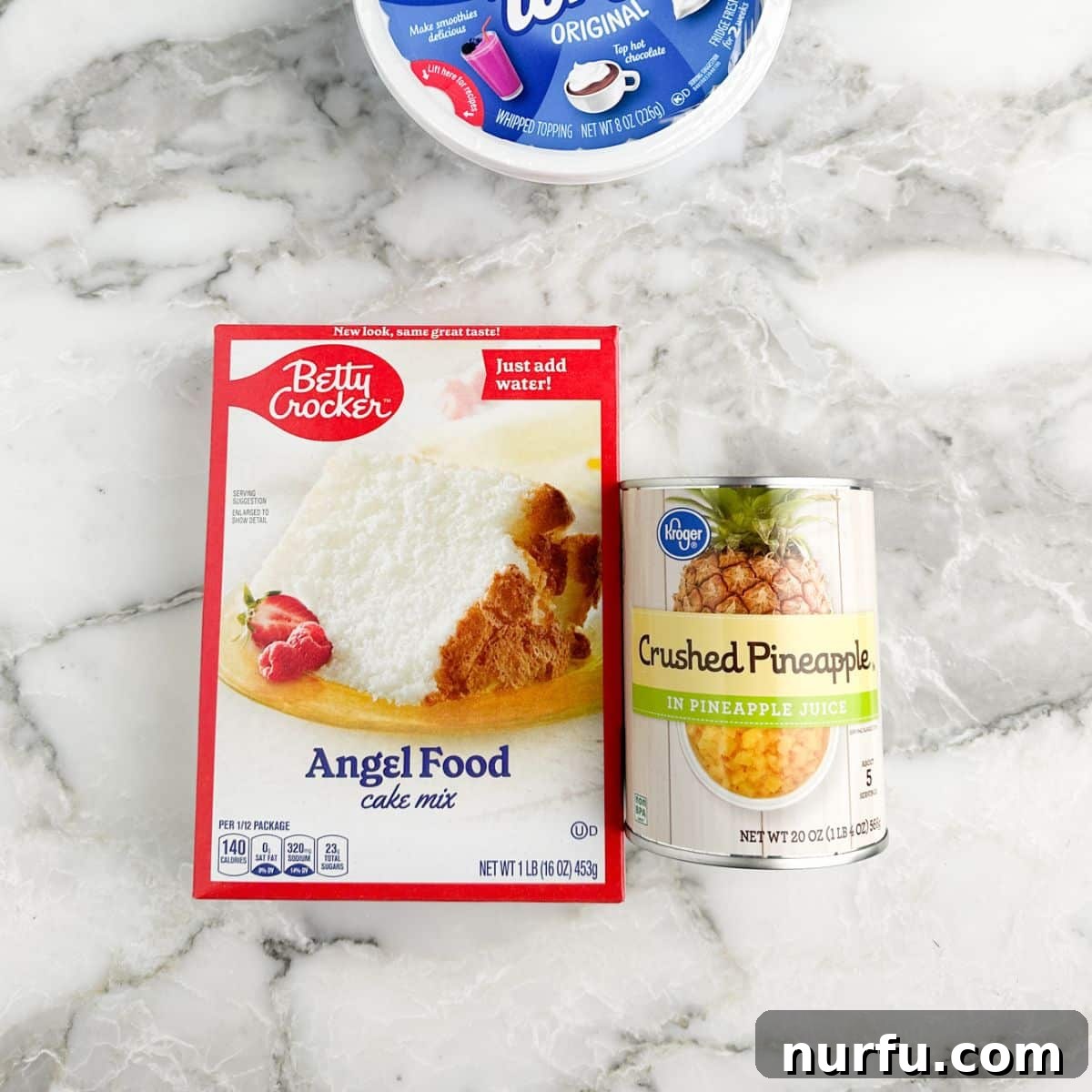 A box of angel food cake mix, a can of crushed pineapple, and a tub of Cool Whip are neatly arranged, showcasing the simple ingredients for the cake.