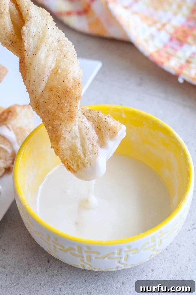 A close-up shot of a cinnamon twist dipped into a bowl of creamy white icing, highlighting its deliciousness.