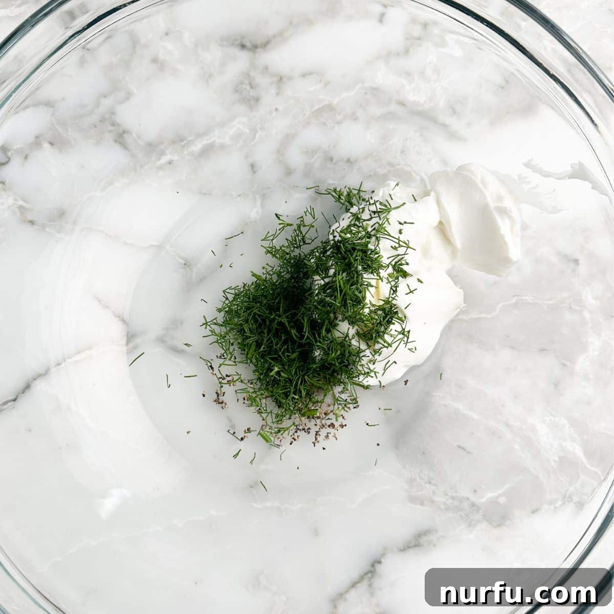 Refreshing Creamy Cucumber Dill Salad 4 A bowl containing a creamy white dressing, freshly chopped green dill, and seasonings, ready for mixing.