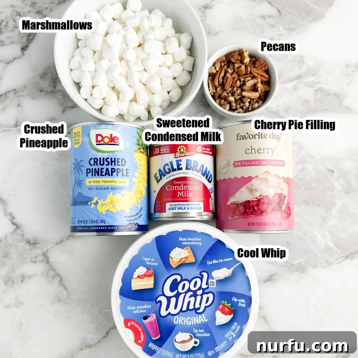 A selection of cherry fluff ingredients including marshmallows, chopped pecans, crushed pineapple, cherry pie filling, condensed milk, and cool whip laid out on a surface.