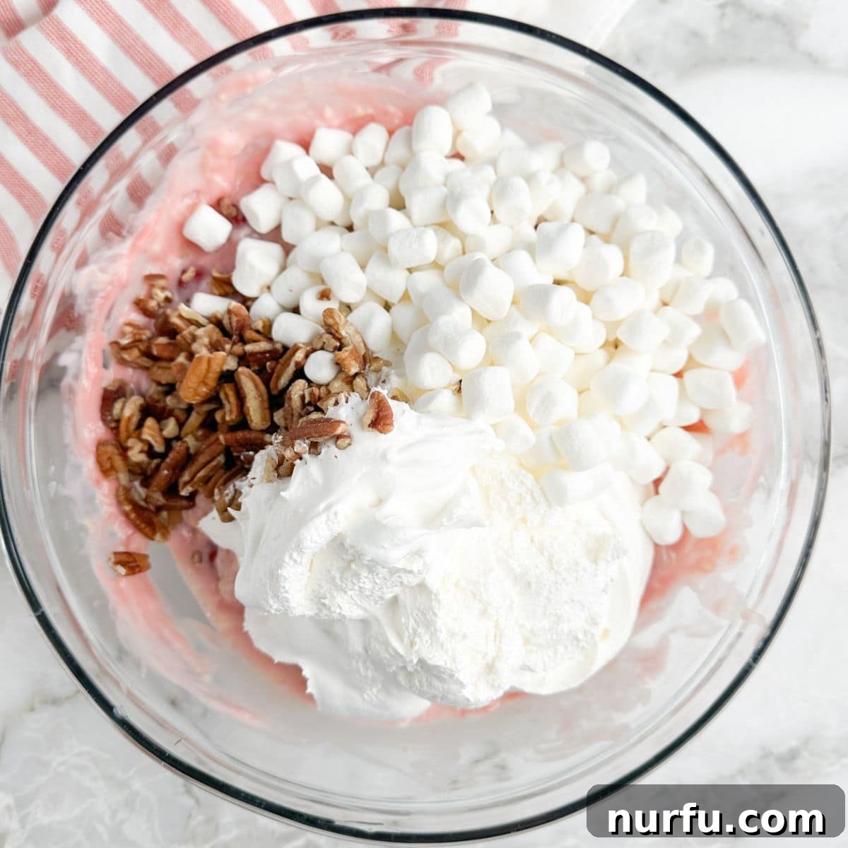 A bowl with mini marshmallows, chopped pecans, and Cool Whip being stirred into the cherry mixture.