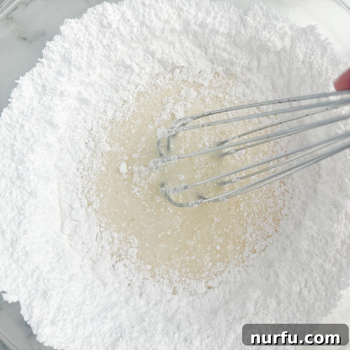 A white bowl containing powdered sugar, with a metal whisk resting inside, signaling the start of the lemon icing preparation.