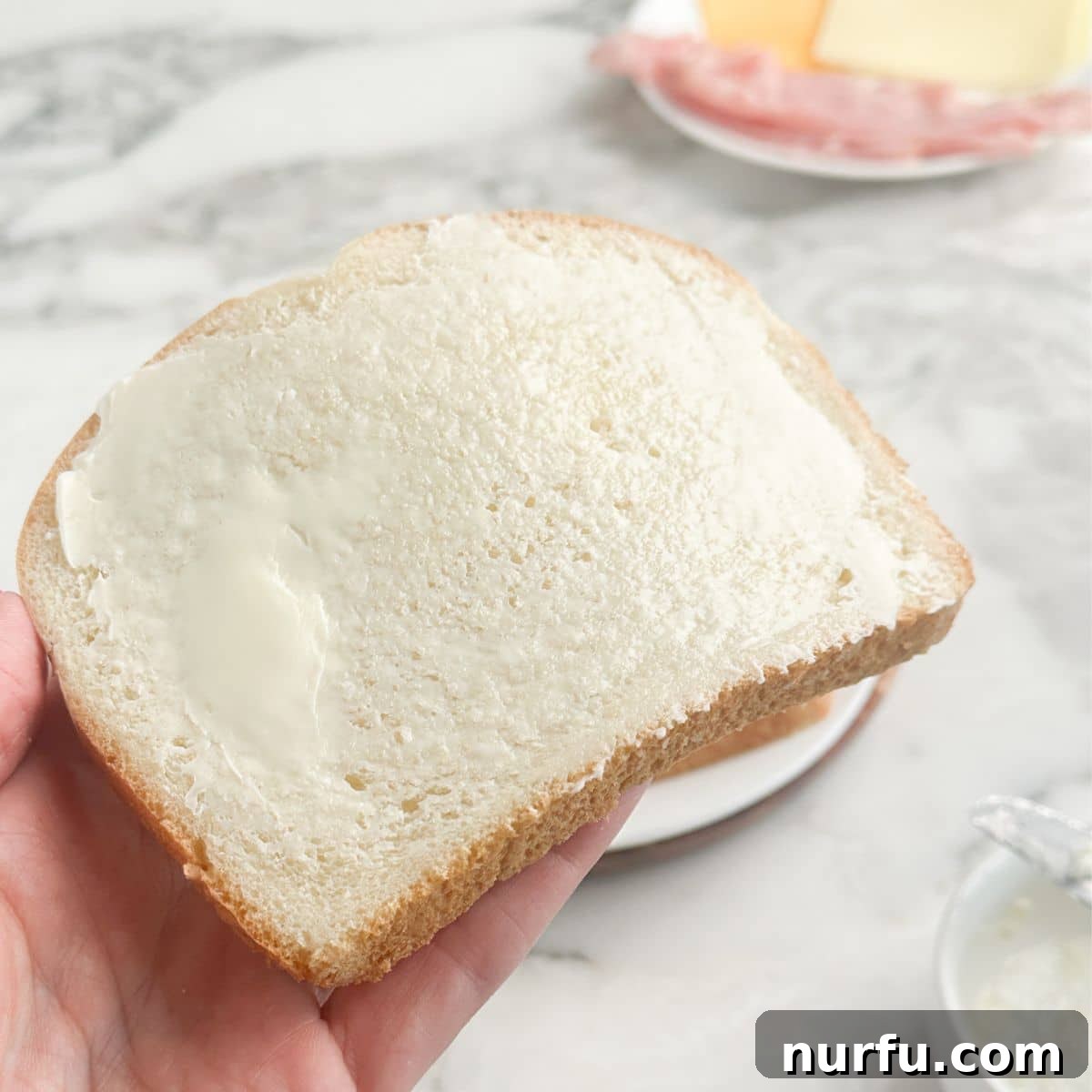 Ultimate Ham and Cheese Melt 4 A hand carefully spreading softened butter onto a slice of sandwich bread, ensuring full coverage to the edges.