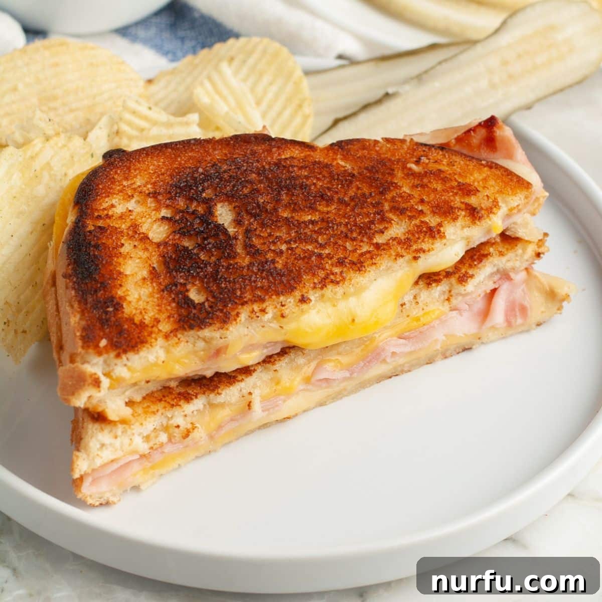 Ultimate Ham and Cheese Melt 9 A freshly cooked grilled ham and cheese sandwich, perfectly sliced, served on a plate with a side of crisp chips and a pickle.