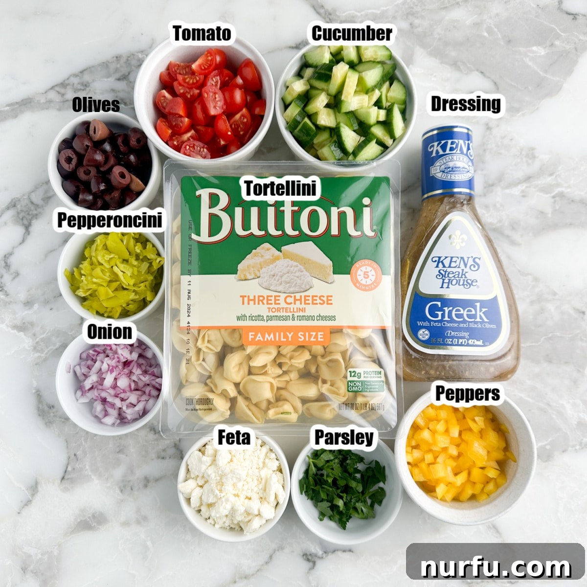 Vibrant Greek Tortellini Pasta Salad 3 Individual bowls containing fresh ingredients for Greek tortellini pasta salad, including tortellini, sliced cucumbers, bell peppers, Kalamata olives, red onion, feta cheese, and a bottle of Greek dressing.