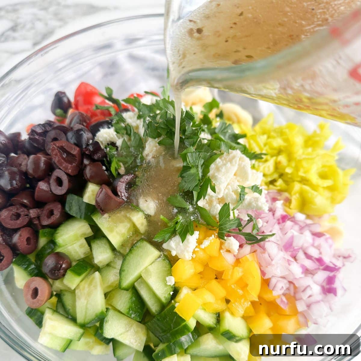 Vibrant Greek Tortellini Pasta Salad 4 A large mixing bowl with cooked tortellini, chopped olives, cucumbers, and red onions, with Greek dressing being poured over the ingredients before tossing.
