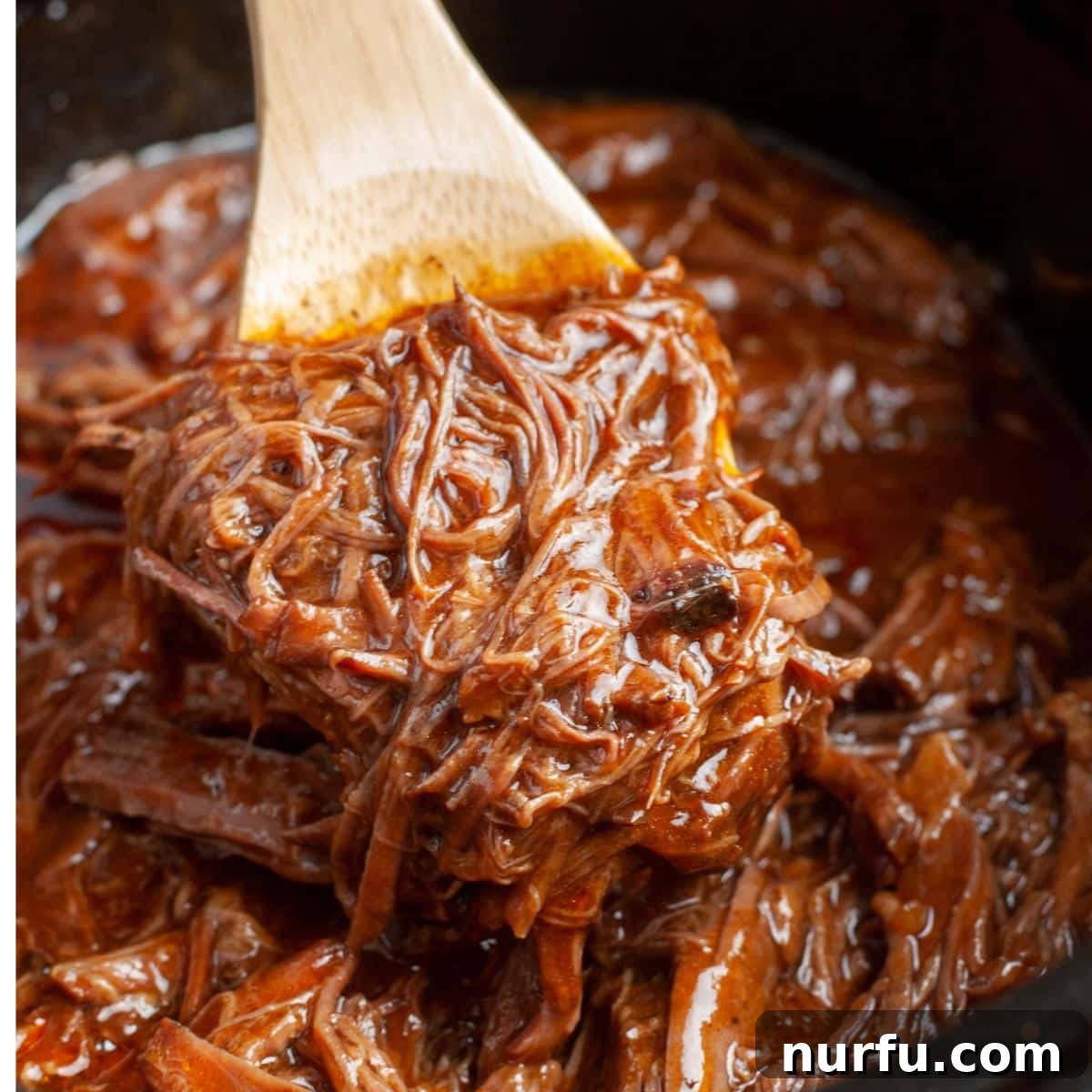 Fall Apart Slow Cooker BBQ Beef 7 A wooden spoon lifting a spoonful of perfectly shredded, dark brown BBQ beef from the slow cooker, demonstrating its tender, fall-apart texture.