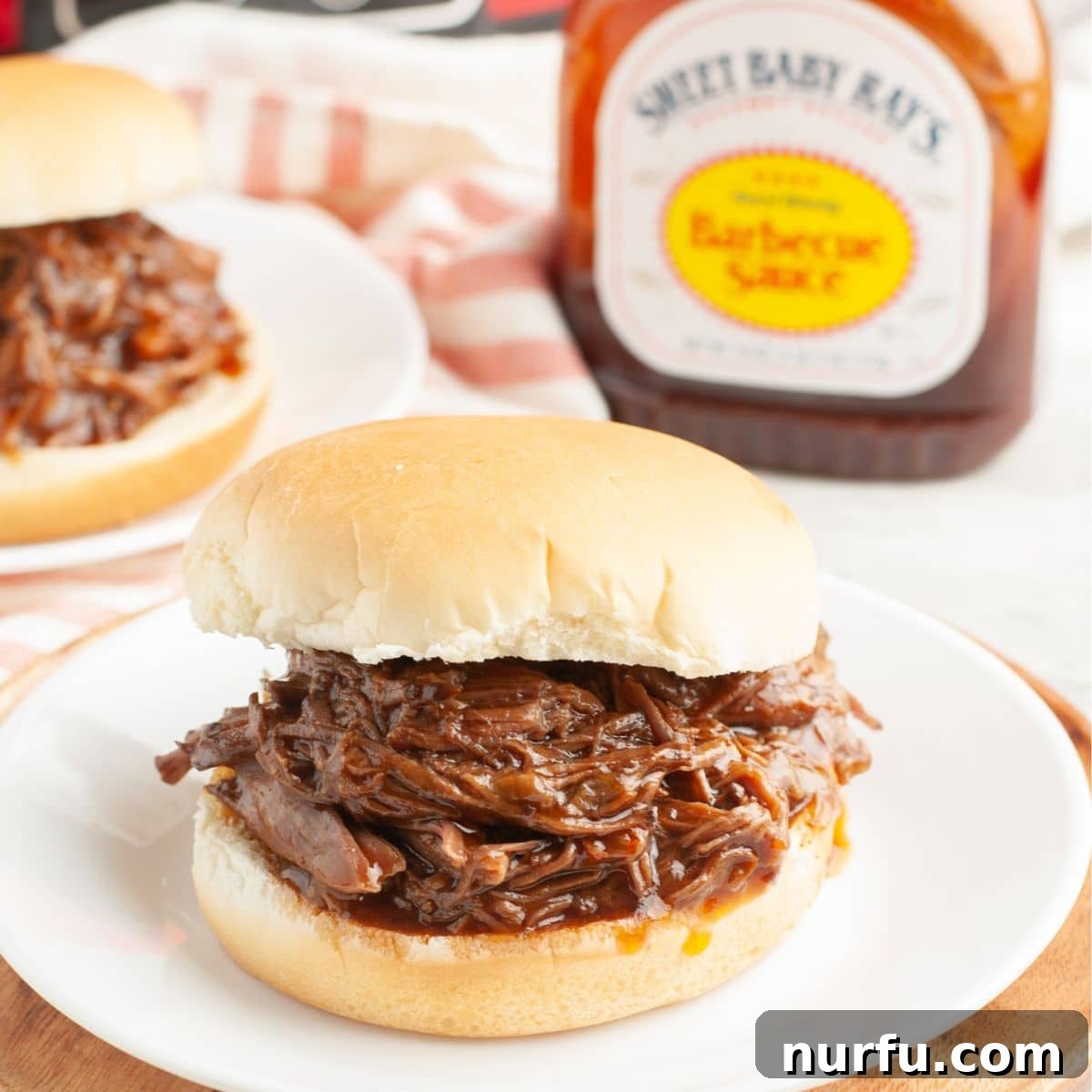 Fall Apart Slow Cooker BBQ Beef 8 A delectable slow cooker BBQ beef sandwich on a white plate, accompanied by a small bottle of barbecue sauce, ready for a flavorful meal.