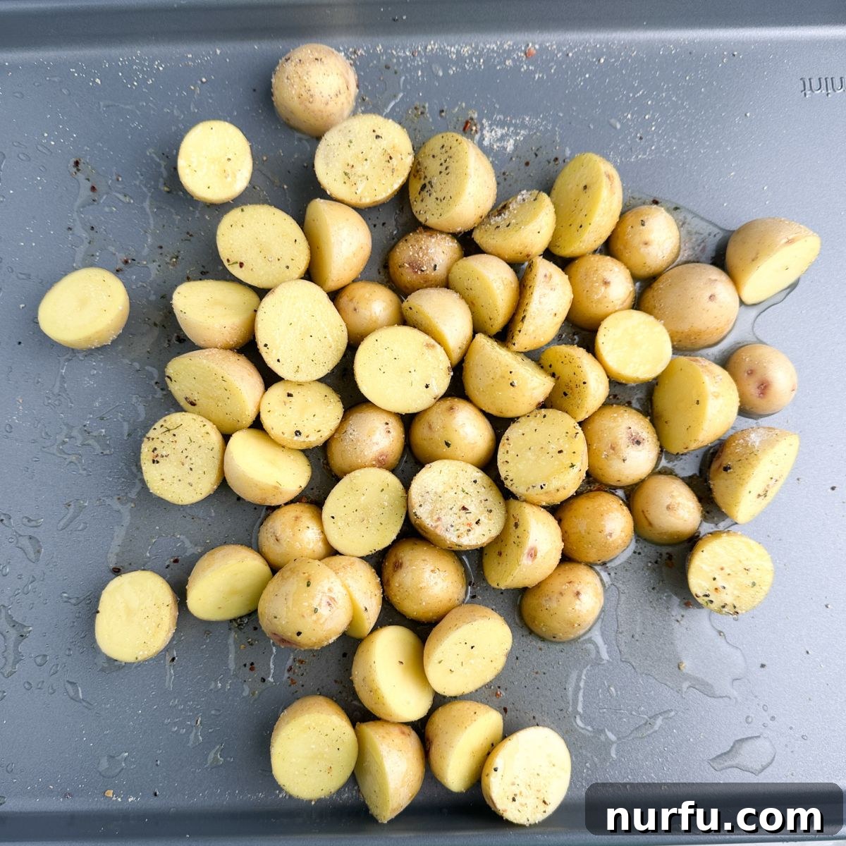 Mini potatoes cut in half on a baking sheet, drizzled with olive oil and sprinkled with seasoning.