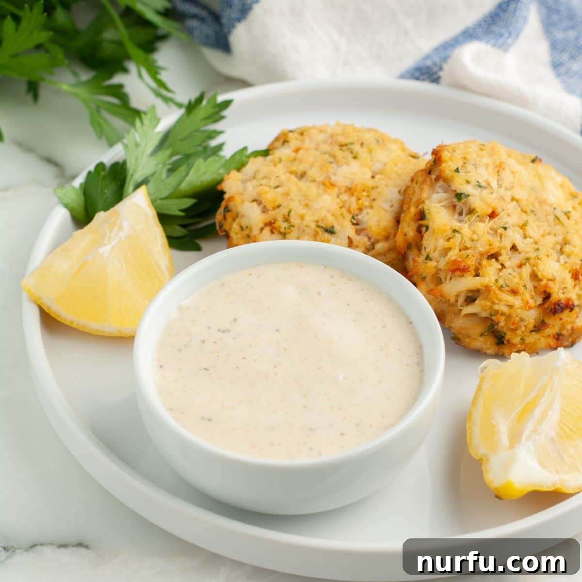 Homemade Crab Cake Sauce Perfection 6 A bowl of creamy Old Bay sauce served alongside perfectly golden-brown crab cakes, garnished with fresh herbs.