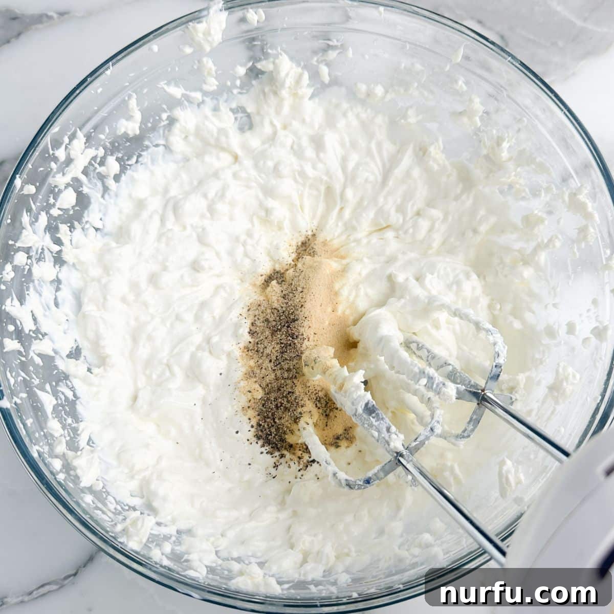 Savory Scallion Dip 4 A mixing bowl filled with beaten softened cream cheese and sour cream, along with various seasonings scattered around, as a hand mixer rests in the bowl after achieving a creamy consistency.
