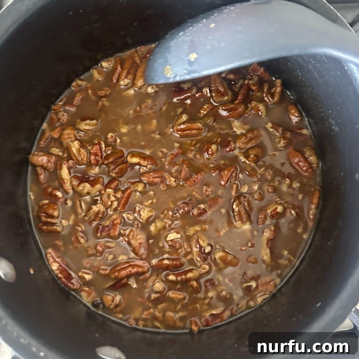 A saucepan on a stovetop, containing a bubbling mixture of butter, brown sugar, and chopped pecans, forming the pecan pie topping.