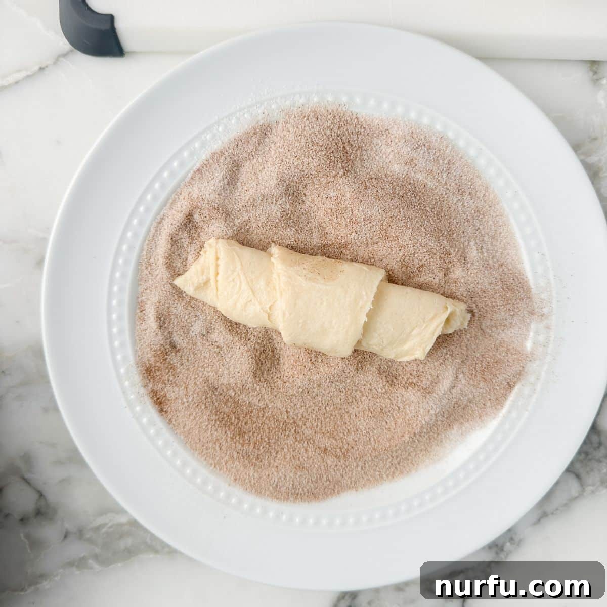 A rolled crescent dough piece being coated in a mixture of cinnamon and sugar spread on a flat plate.
