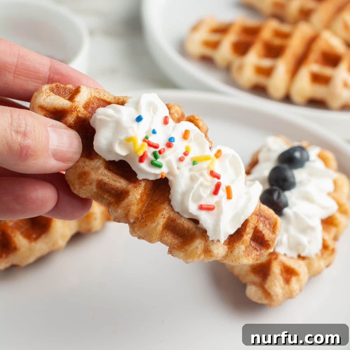 A hand holding a perfectly cooked croffle, topped with whipped cream and colorful sprinkles, ready to be enjoyed.