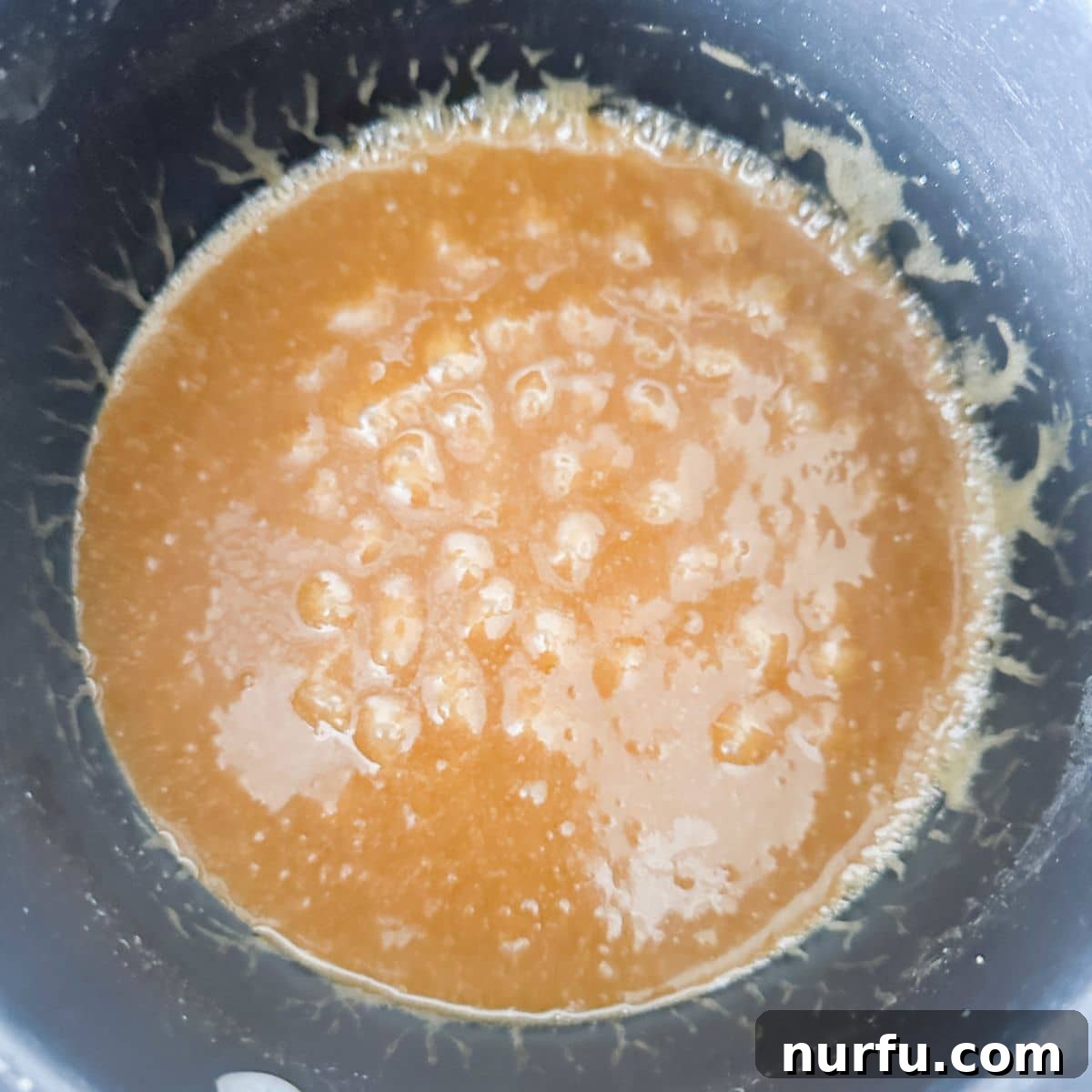 A medium saucepan on a stovetop, filled with a bubbling, golden-brown butter toffee sauce, showing the rich texture of the caramel as it cooks.